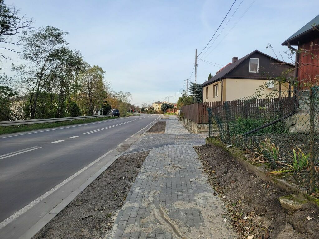 
                                                    Obraz przedstawia nowo wybudowany chodnik wzdłuż miejskiej drogi. Po jednej stronie chodnika znajduje się metalowe ogrodzenie, a po drugiej domy i drzewa.
                                                