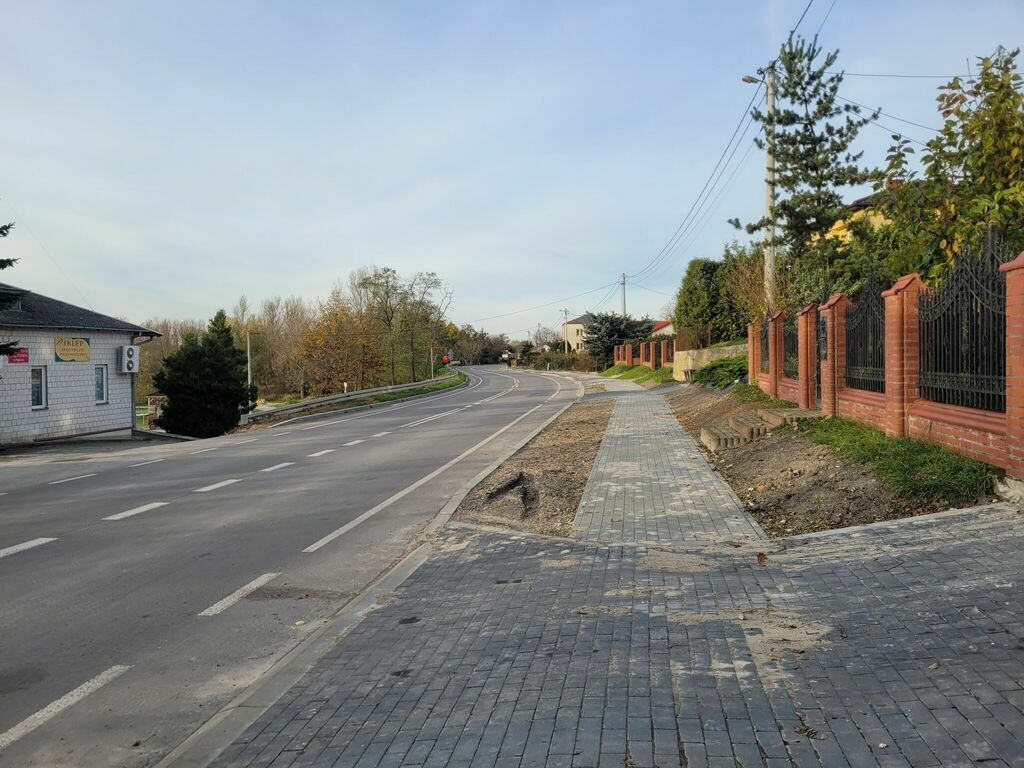 
                                                    Nowo wybudowany chodnik wzdłuż ulicy, po prawej czerwone ogrodzenie i zieleń, po lewej budynek i znak drogowy.
                                                