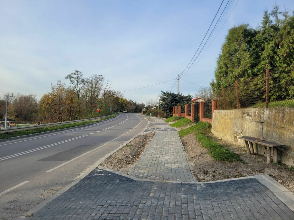 
                                                    Nowo wybudowany chodnik z kostki brukowej wzdłuż krętej drogi. Po prawej stronie ogrodzenie i ławka, po lewej drzewa i krzewy.
                                                