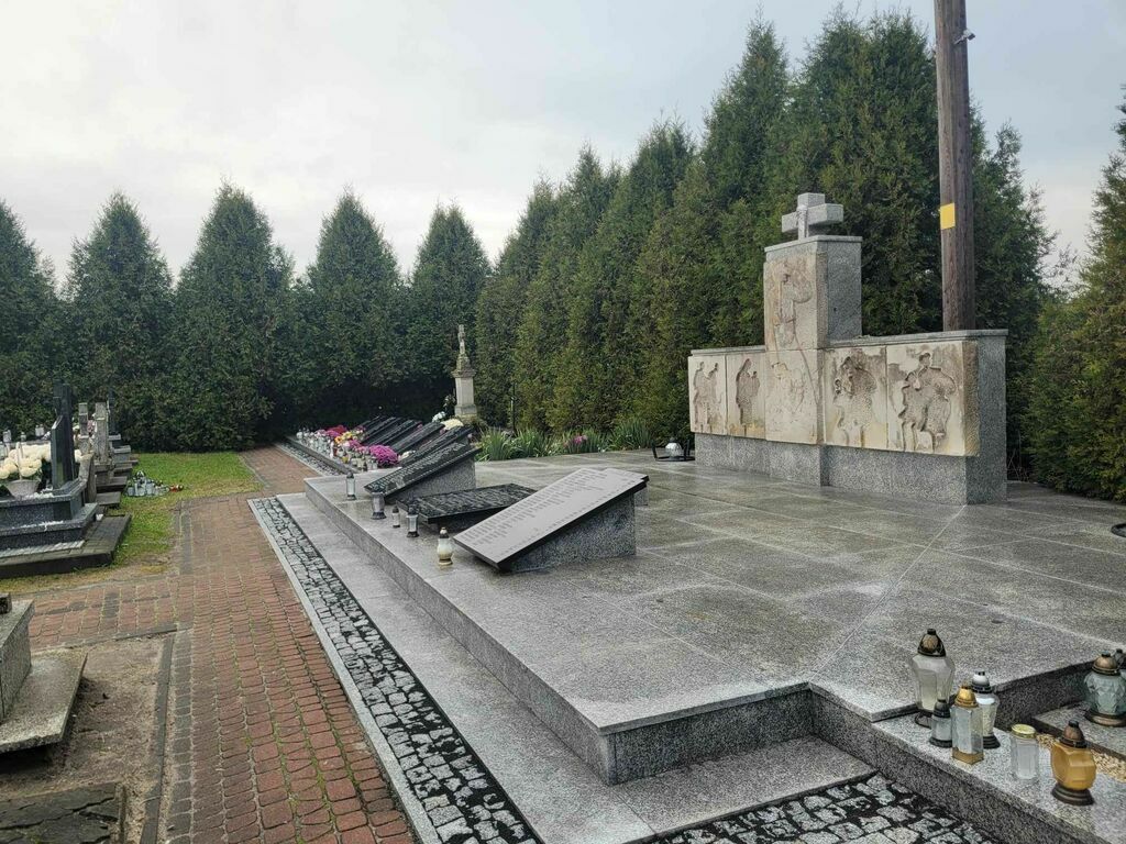 
                                                    Zdjęcie przedstawia odnowiony cmentarz z mogiłą zbiorową, granitowymi płytami, krzyżem i wieńcami na tle drzew.
                                                