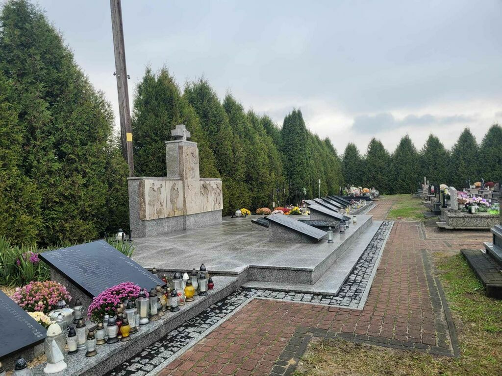 
                                                    Zdjęcie przedstawia odnowiony cmentarz z kamiennym pomnikiem centralnym i nawierzchnią z kostki brukowej, otoczony zniczami i kwiatami na tle zielonych drzew.
                                                