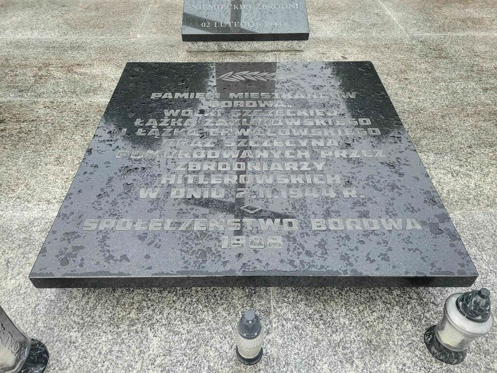 
                                                    Pomnik z czarnego kamienia z wygrawerowanym tekstem poświęcony ofiarom pacyfikacji, otoczony przez znicze.
                                                