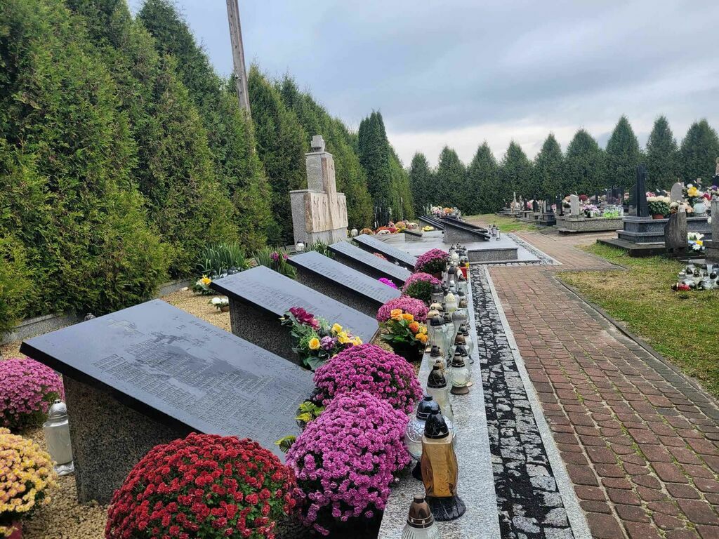 
                                                    Zdjęcie przedstawia zadbany cmentarz z kamiennymi płyty z inskrypcjami, położonymi na ziemi, otoczonymi kwiatami i zniczami, z pomnikiem w tle.
                                                