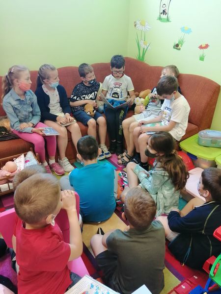 
                                                    Zdjęcie przedstawia kilkoro dzieci siedzących na kanapie wśród nich jest chłopiec czytający książkę oraz część dzieci siedzących na dywanie i słuchających kolegi.
                                                