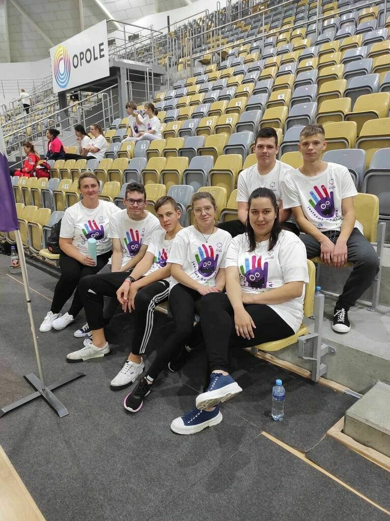 
                                                    Grupa osób siedzi na trybunach stadionu, ubrani w białe koszulki z logo Igrzysk bez Barier.
                                                