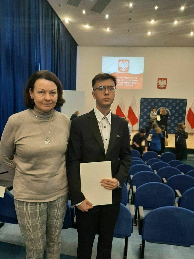 
                                                    Młody mężczyzna w garniturze stoi obok kobiety w swetrze. Trzymają dyplom. W tle sala z rzędami foteli i ekran z herbem Polski.
                                                