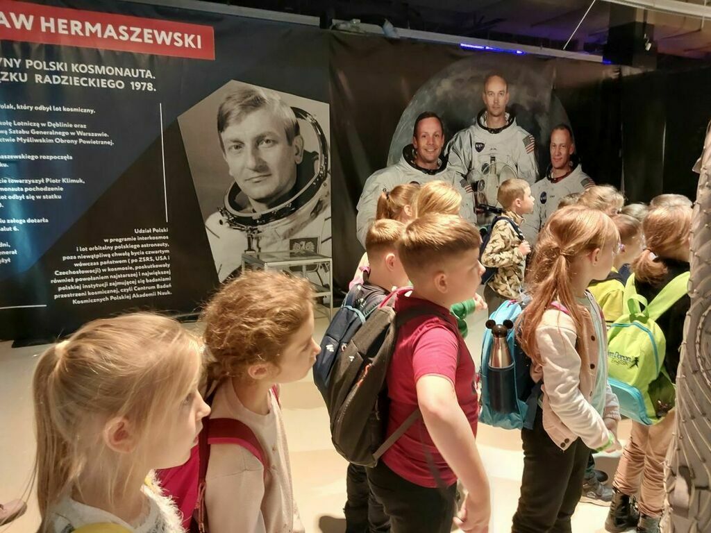 
                                                    Grupa dzieci stoi przed ekspozycją kosmiczną z figurą astronauty, obok informacje o pierwszym polskim kosmonaucie, Mirosławie Hermaszewskim.
                                                