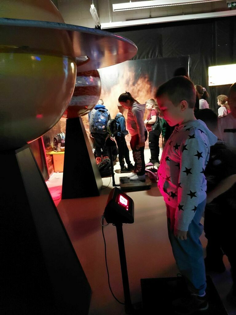 
                                                    Chłopiec obserwuje eksponaty na wystawie kosmicznej, w tle inne dzieci i modele planet, atmosfera naukowa i edukacyjna.
                                                