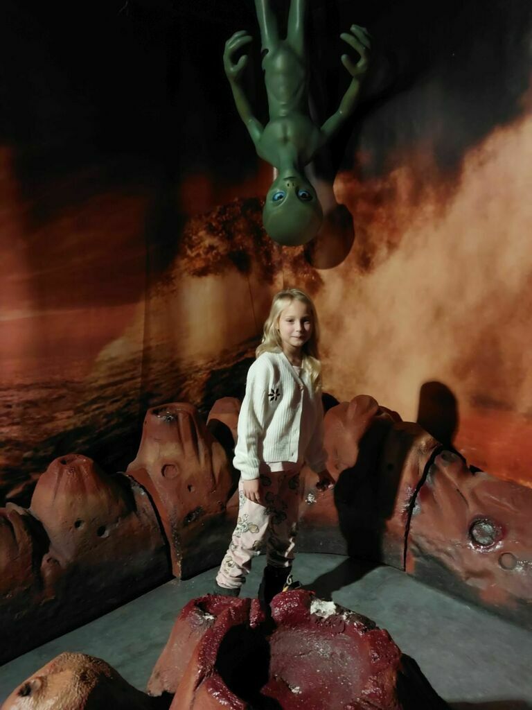 
                                                    Dziewczynka stoi na wystawie kosmicznej, w tle pomarańczowy żar z wulkanu i wiszący zielony kosmita.
                                                