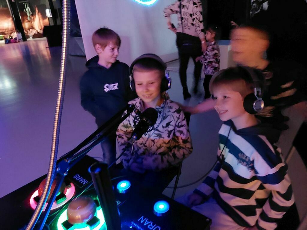 
                                                    Dzieci korzystają z interaktywnej instalacji z przyciskami i słuchawkami na wystawie KOSMOPARK, w tle inne dzieci się bawią.
                                                