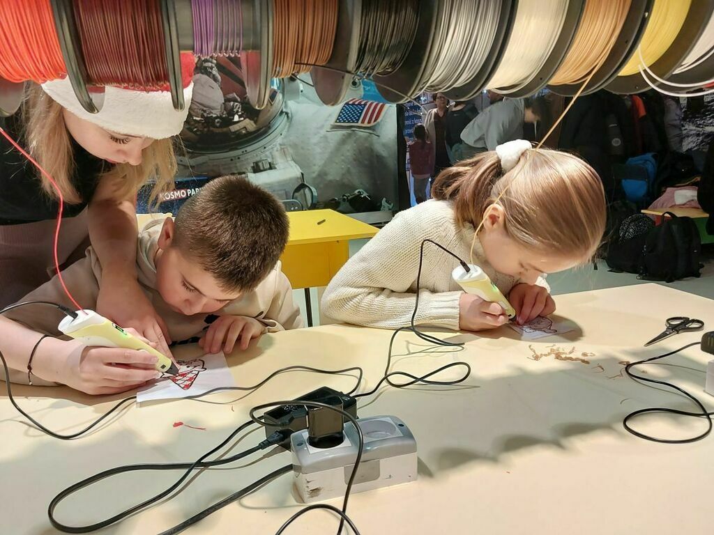 
                                                    Dzieci uczestniczą w warsztacie, używając lutownic do połączeń elektrycznych; w tle widoczne są kolorowe rolki z przewodami.
                                                