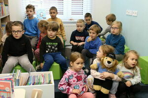 Grupa przedszkolaków siedzi w pokoju z książkami; niektóre dzieci trzymają maskotkę, wszystkie skupione na czymś poza kadr.