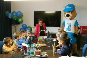 Grupa przedszkolaków siedzi przy stole z przekąskami, na którym widać również zielono-niebieskie balony. Po prawej stronie stoi maskotka, a po lewej dorosła kobieta obserwuje dzieci.