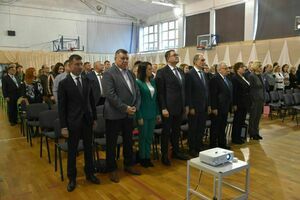 Grupa ludzi stoi w rządku w sali gimnastycznej przekształconej na salę ceremonii, zwróceni w jednym kierunku, w tle widoczne zasłony i dekoracje.