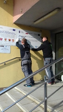 
                                                    Dwie osoby przyczepiające tablice informacyjne
                                                