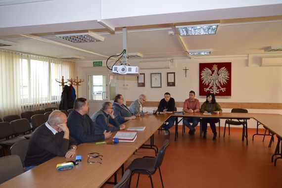 
                                                    Grupa ludzi siedzących w ławkach na tle godła
                                                