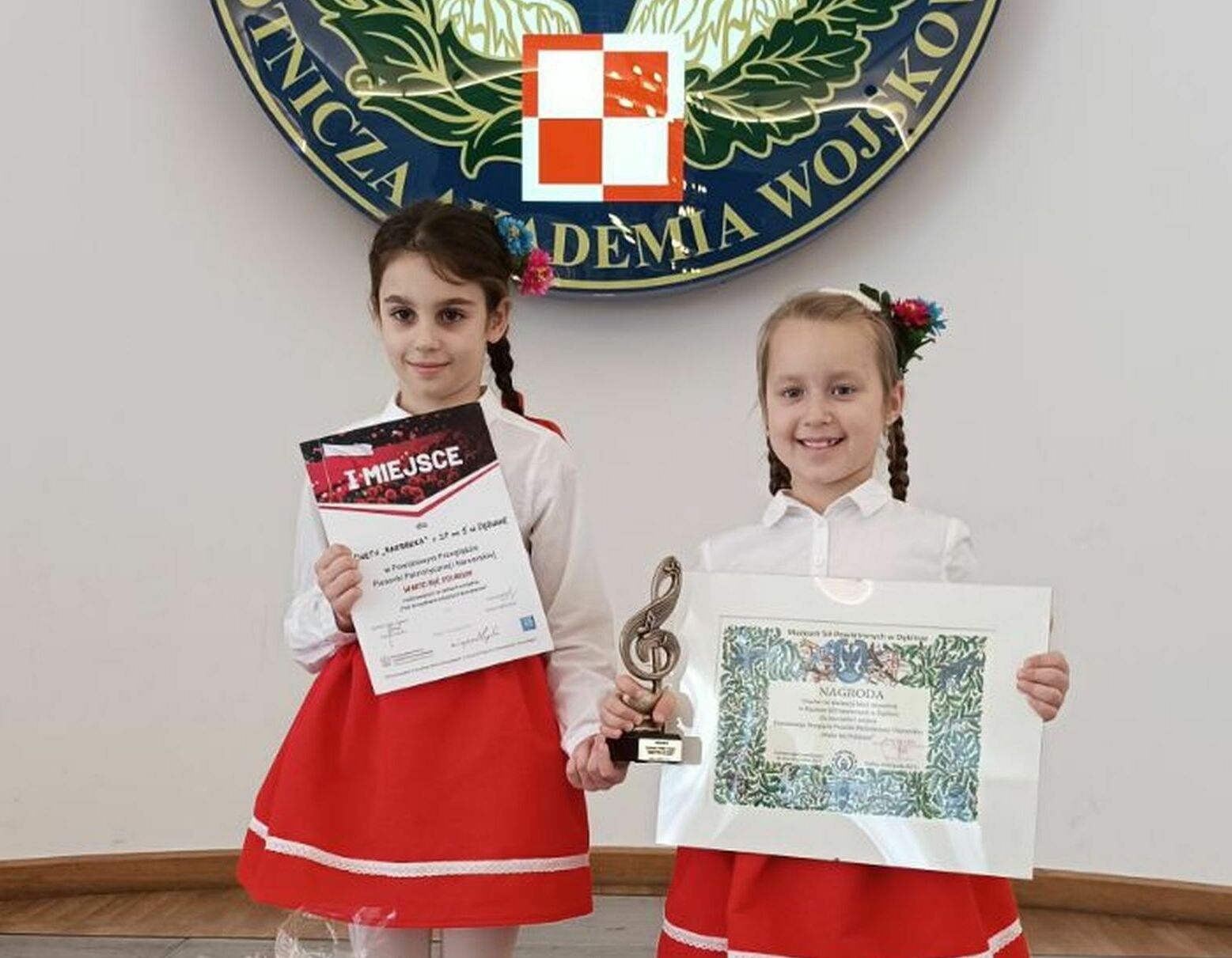 Dwie młode dziewczynki w czerwonych spódnicach i białych bluzkach trzymają dyplomy i statuetkę. Stoją przed emblematem z orłem.