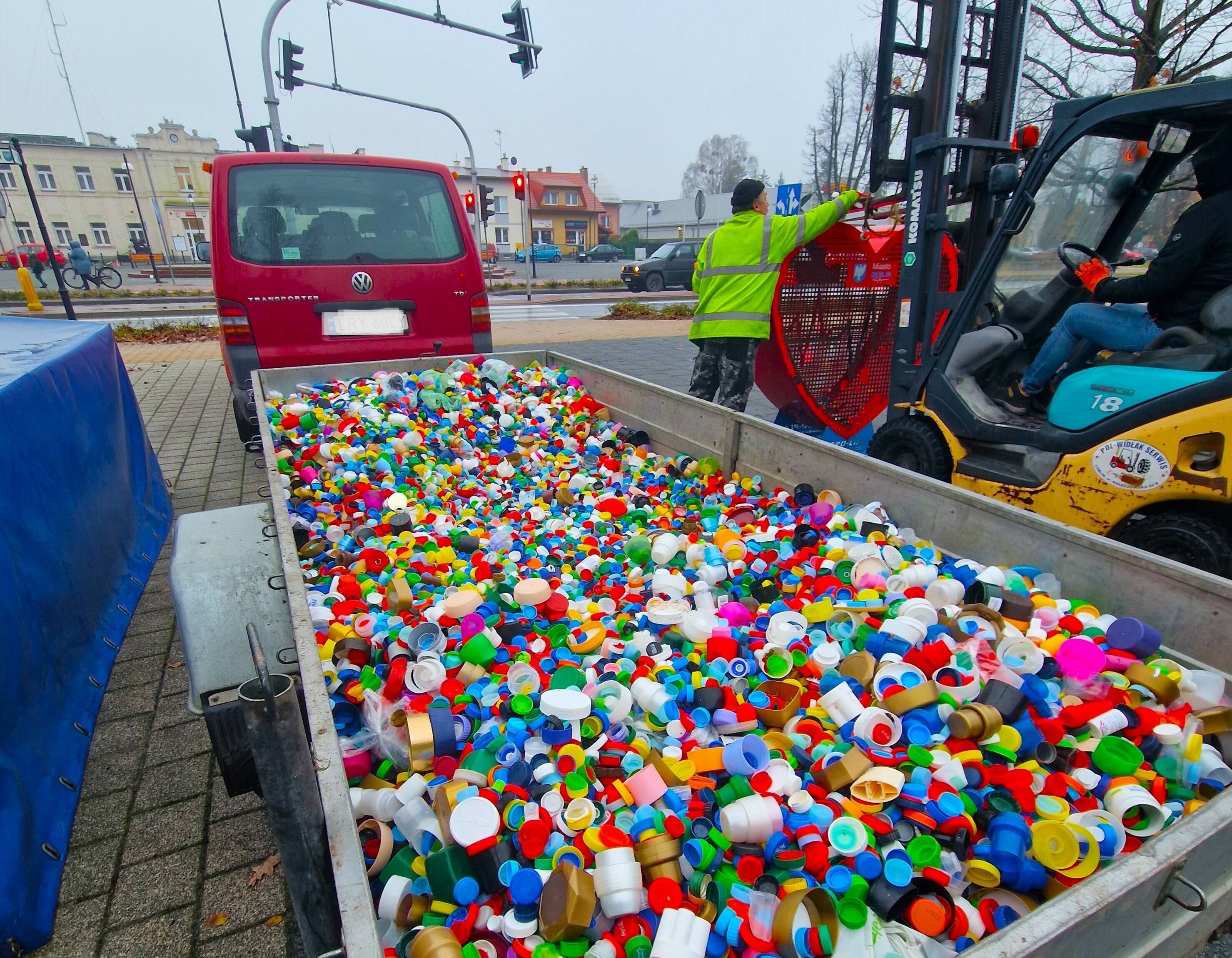Przyczepa wypełniona kolorowymi plastikowymi nakrętkami; pracownik komunalny w odblaskowej kamizelce; miejski pejzaż w tle.