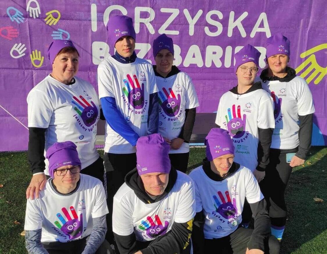 Grupa ośmiu uśmiechniętych osób w fioletowych czapkach i białych koszulkach z różnokolorowymi odciskami dłoni. Stoją przed banerem z napisem "Igrzyska Integracji".