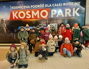 Grupa dzieci w kolorowych czapkach stoi przed banerem "KOSMO PARK" w środku centrum handlowego.