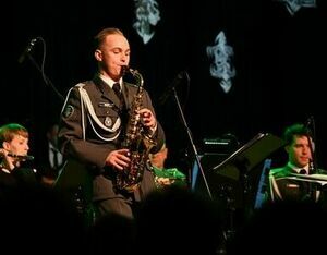 Mężczyzna w mundurze wojskowym gra na saksofonie altowym na scenie, w tle muzycy z nutami.
