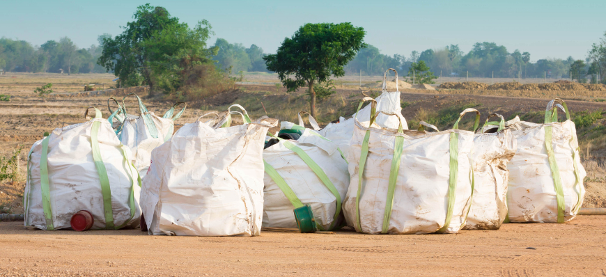 Duże, białe worki typu big bag wypełnione śmieciami, ustawione w rzędzie na obrzeżach pola z brązową ziemią w tle.