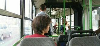 Wnętrze zielonego autobusu komunikacji miejskiej z siedzącymi i stojącymi pasażerami, jadącego przez miejską ulicę w ciągu dnia.