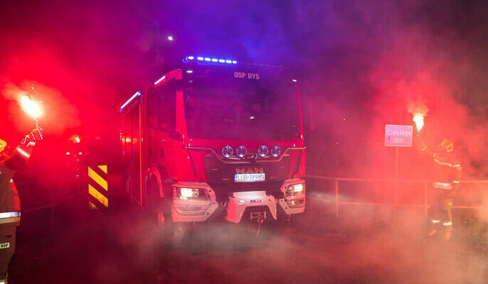 Samochód strażacki, a po jego stronach strażacy z zapalonymi flarami