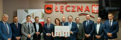 Grupa jedenastu osób, mężczyzn i kobiet, w formalnym ubraniu stojących i uśmiechających się do kamery, trzymających wspólnie dużą czekową tablicę, na tle logotypu firmy.