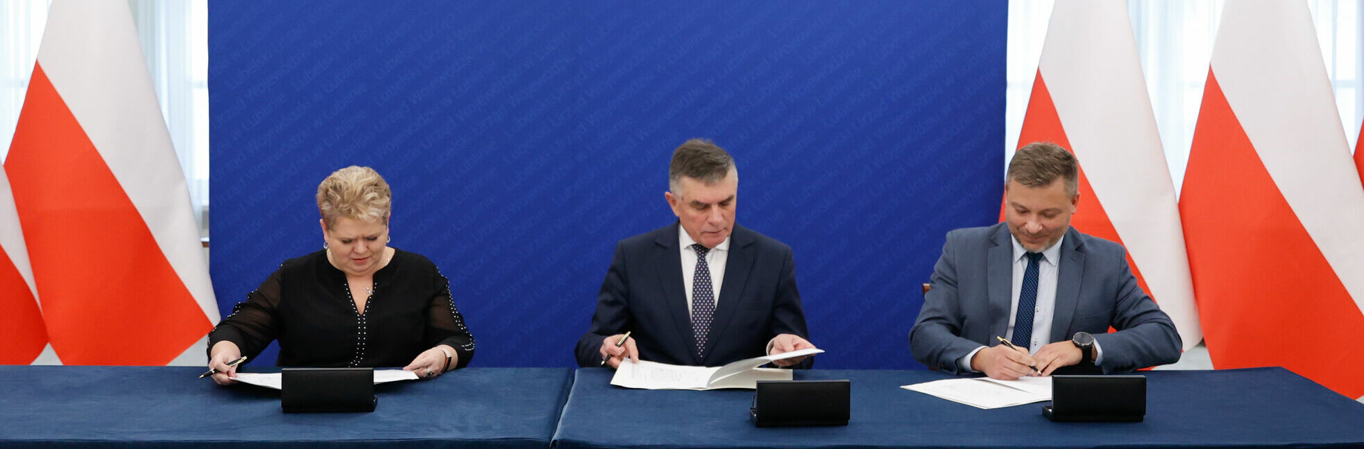 Trzy osoby siedzą przy stole z dokumentami na tle flag Polski oraz niebieskiej ściany z białymi pasami, podpisując oficjalne dokumenty.