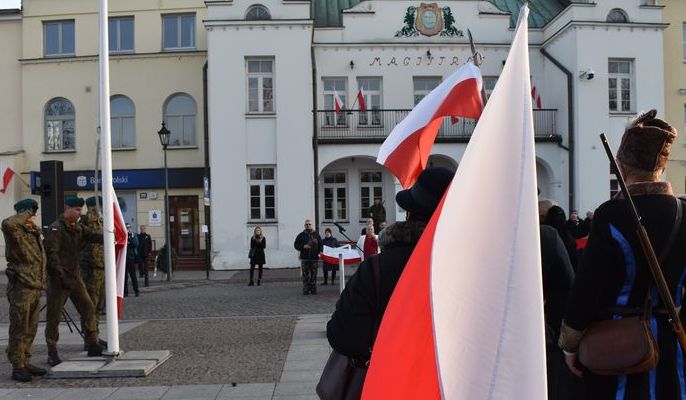 Żołnierze wciągają flagę Polski na maszt, obok stoją ludzie trzymający flagi narodowe