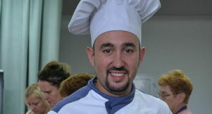 Mężczyzna w białym uniformie kucharskim i czapce uśmiecha się do kamery, w tle rozmyci ludzie.