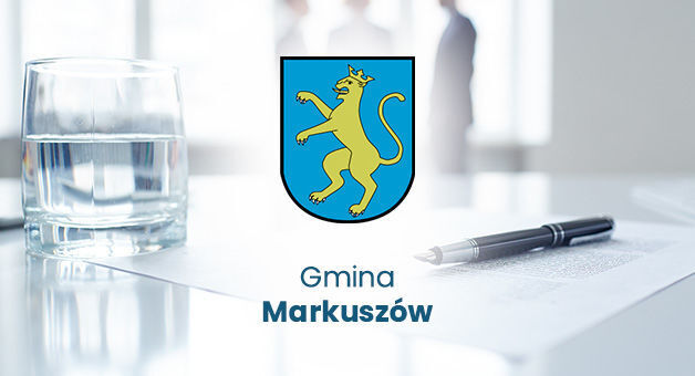 Na zdjęciu znajduje się logo gminy Markuszów - błękitna tarcza herbowa z żółtym lwem w środku. W tle rozmyte biuro, szklanka wody i długopis leżą na stole.