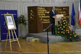 Mężczyzna stoi na podium w ozdobionej sali z kwiatami i plakatem o edukacji. Obok podium portret na sztaludze.