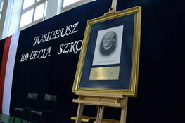 Obraz przedstawiający portret osoby w złotej ramie na sztaludze, z plakietką poniżej, na tle banera z napisem "JUBILEUSZ 100-LECIA SZKOŁY".