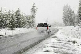 Samochód jedzie śnieżną, zakrętową drogą otoczoną przez zasypane śniegiem drzewa iglaste. Pogoda wydaje się być zamglona z powodu padającego śniegu.