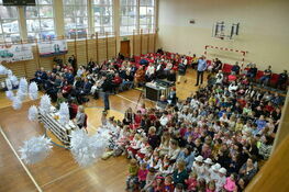 Widok z góry na salę gimnastyczną z grupą ludzi siedzącą na krzesłach i dzieci siedzące na podłodze, zdobione balony, scenariusz wydarzenia publicznego.