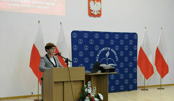 Osoba stoi za mównicą w sali z polskimi flagami, logo MEN i napisem "Konferencja Ministerstwa Edukacji Narodowej".