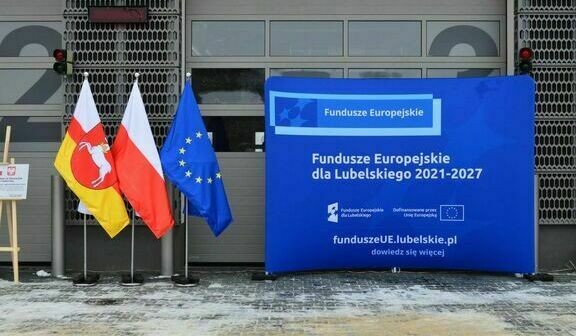 Niebieski baner z napisem "Fundusze Europejskie dla Lubelskiego 2021-2027" przed budynkiem, obok flagi Unii Europejskiej, Polski i z herbem Lublina.