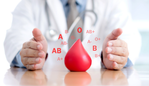 Lekarz w białym fartuchu z fonendoskopem, wyraźnie trzymający czerwoną, kropelkowatą figurę, a w tle różne grupy krwi.