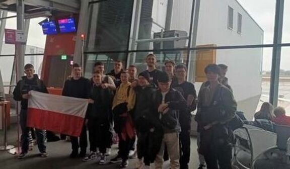 Grupa młodych ludzi stoi razem w hali lotniska, trzymając polską flagę, wyglądając na podekscytowanych, możliwe, że przed podróżą.