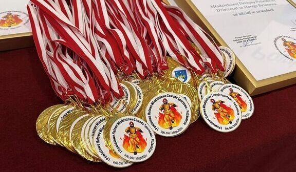Złote medale z czerwono-białymi wstążkami i emblematem leżą na czerwonym materiale obok certyfikatów.