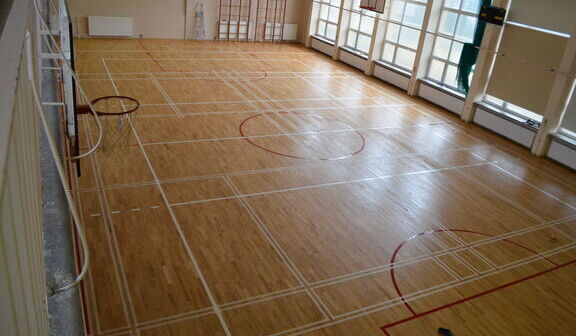 Drewniana podłoga sali gimnastycznej z białymi i czerwonymi liniami oznaczającymi boisko do koszykówki i inne obszary do gier zespołowych.