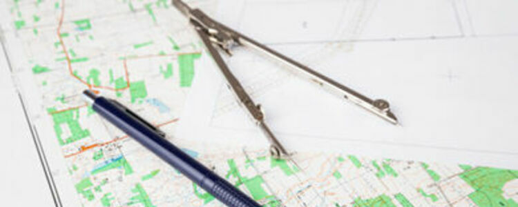 Cyrkiel i długopis leżące na papierze z nieczytelnymi technicznymi rysunkami, zapewne planami lub mapą.