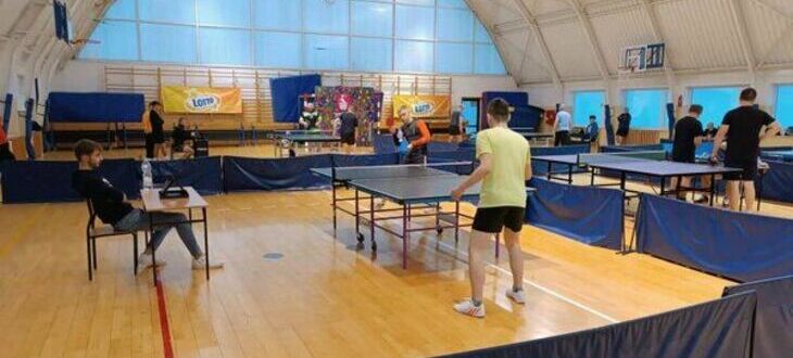 Sala gimnastyczna z kilkoma stołami do tenisa stołowego, gdzie ludzie grają w ping-ponga. Na przedzie mężczyzna czeka na serwis przeciwnika.
