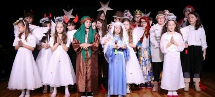 Grupa dzieci przebranych w kostiumy na przedstawienie, w tym anioły, pasterze i Mikołaje, na scenie.