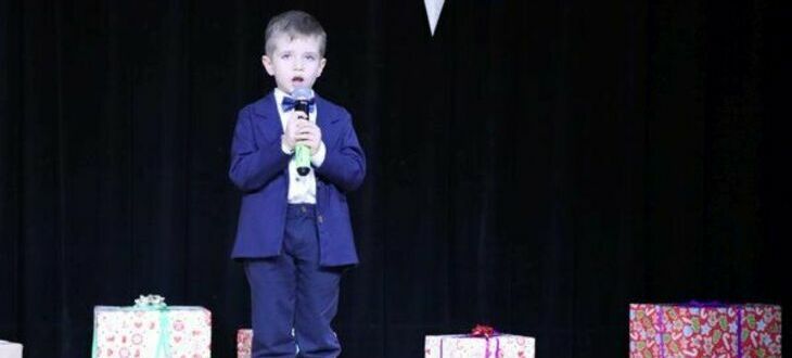Chłopiec w eleganckim garniturze stoi na scenie trzymając mikrofon, w tle widoczne są kolorowe pudełka prezentów.