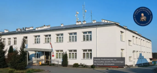 Biały budynek szkoły z niebieskimi akcentami i flagą Polski przy wejściu. Na fasadzie znajduje się tablica z nazwą szkoły.