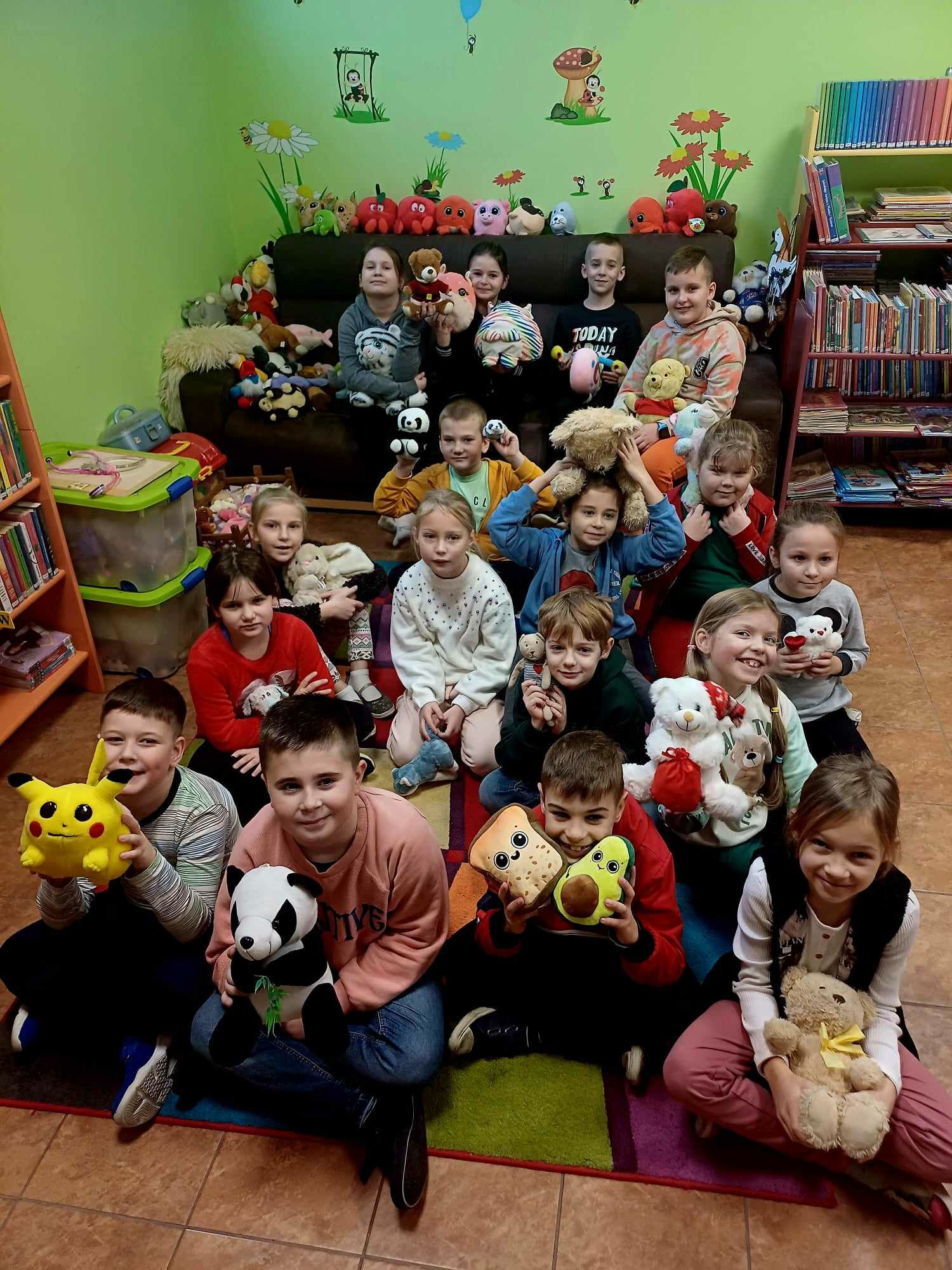 Zdjęcie przedstawia grupę dzieci, które siedzą na kolorowym dywanie, a w rękach trzymają pluszowe maskotki.