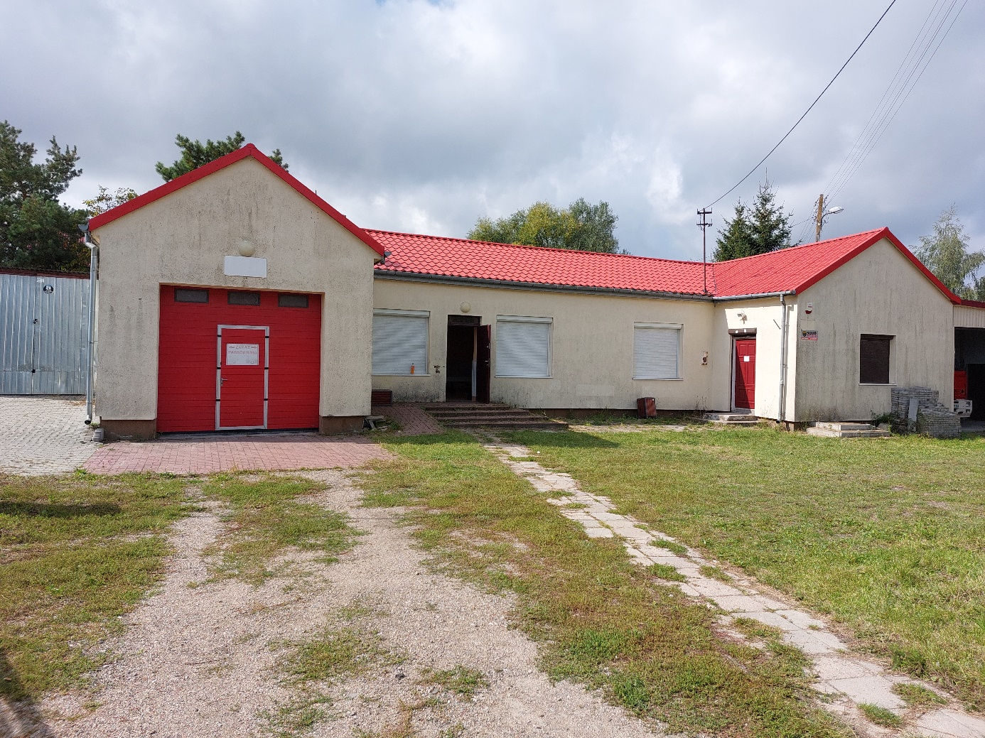 Budynek z jasnej cegły z czerwonym dachem, posiadający czerwone drzwi garażowe i białe wejście, znajdujący się przy żwirowej drodze i trawniku.